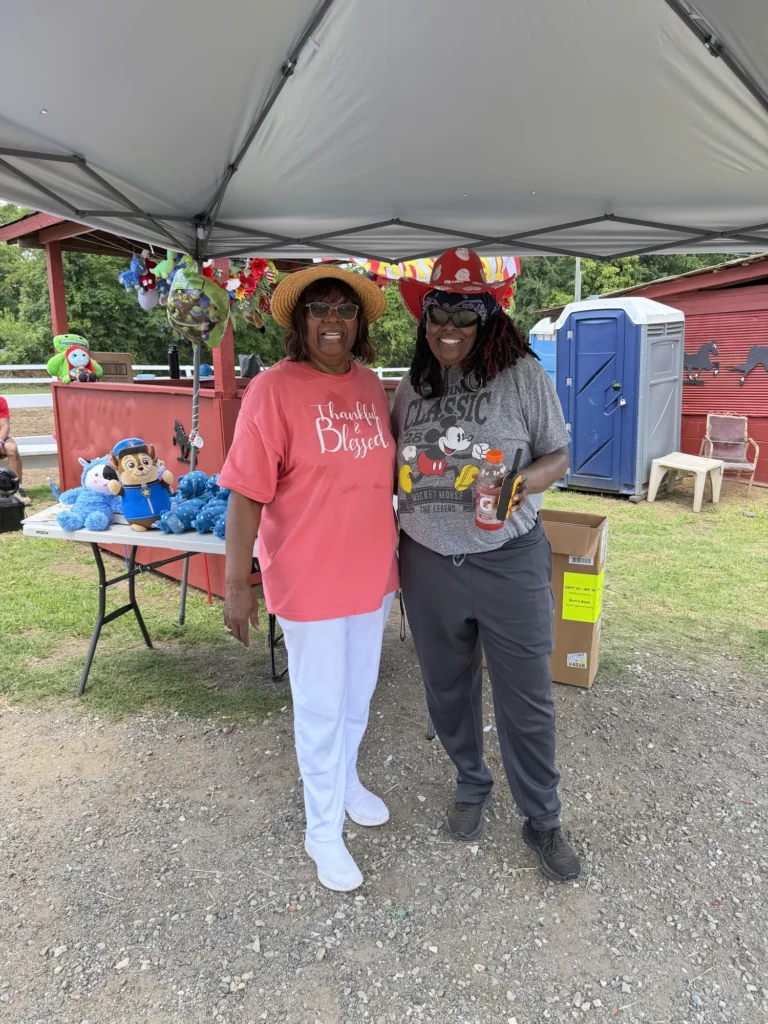 Two volunteers smile beside the carnival prize table stocked with stuffed animals