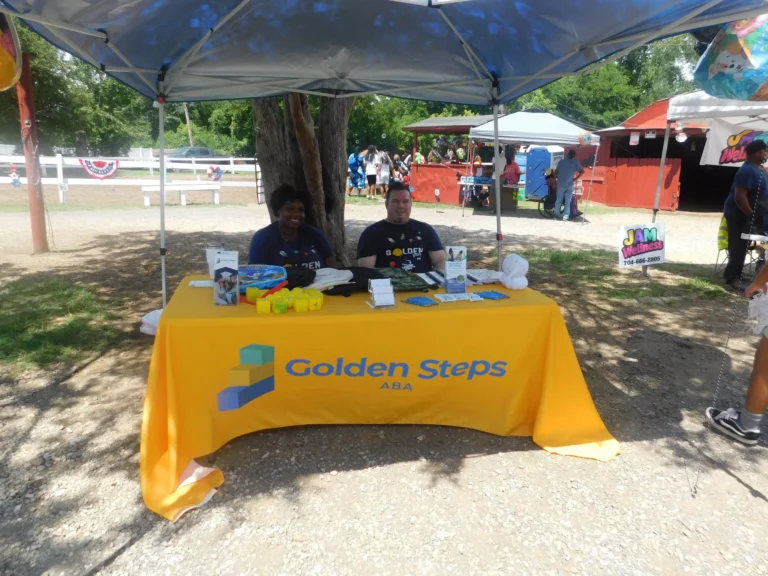 Two volunteers staff the Golden Steps ABA table under a shady canopy