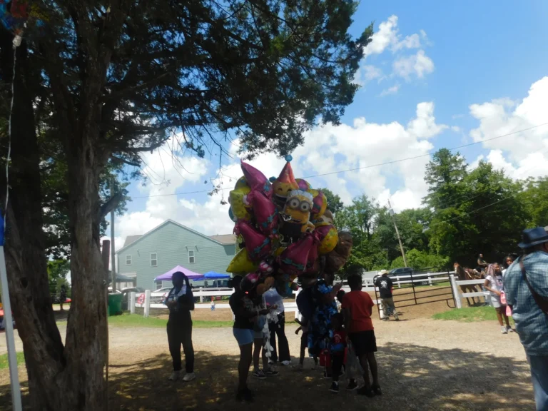 A bunch of character balloons floats above families gathered under the shade trees