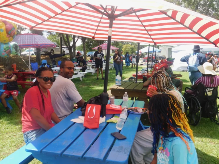 Families share a meal at a picnic table under a striped umbrella