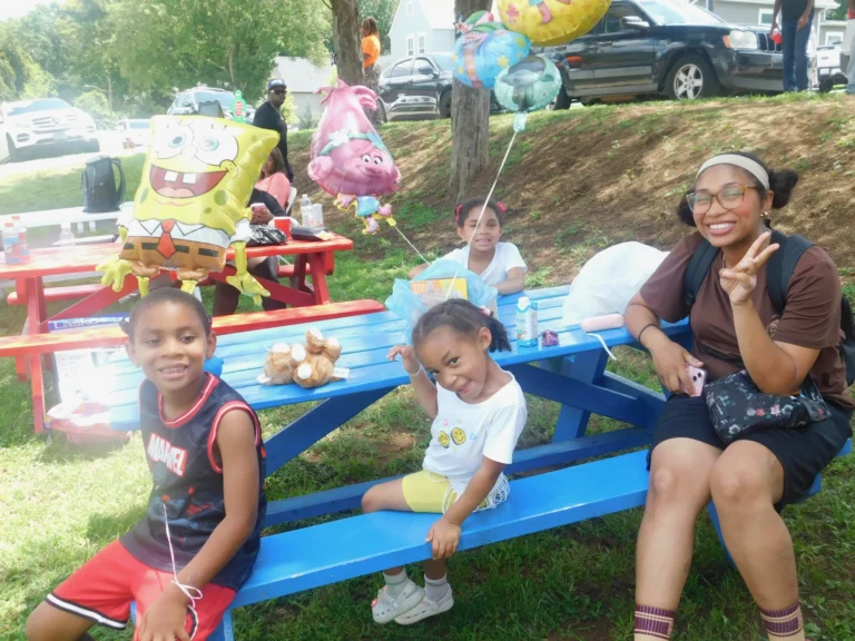 A family gathers at a picnic table with balloons and carnival prizes