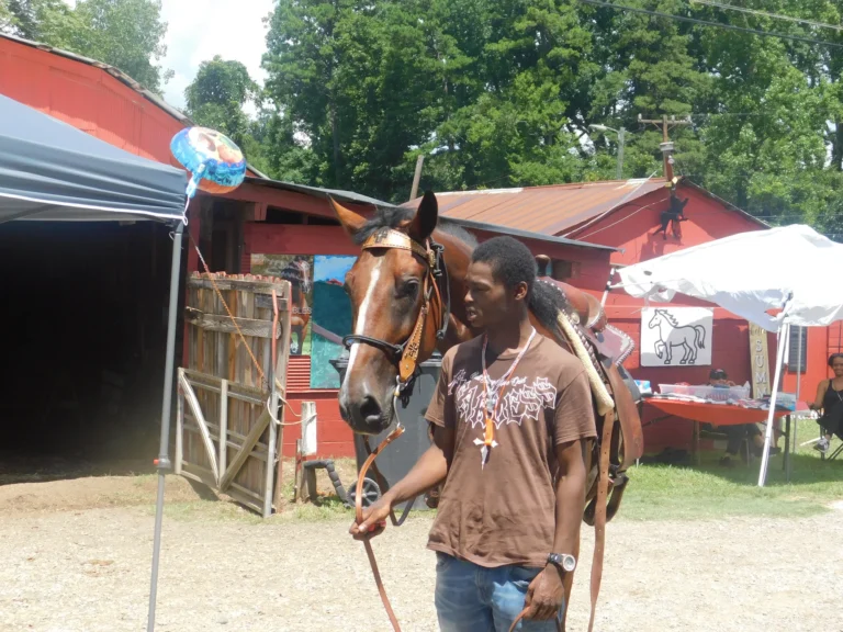 A handler leads a saddled horse past the red barn