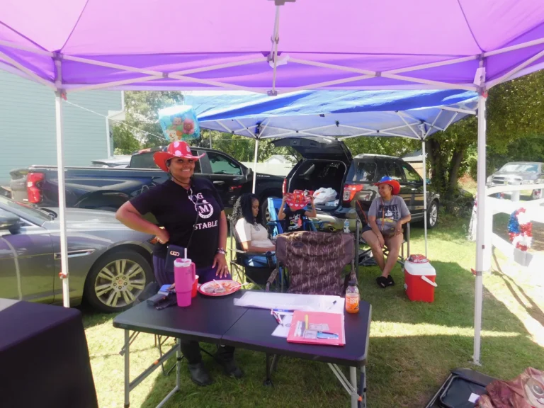 A volunteer in a carnival hat staffs a table under a purple canopy
